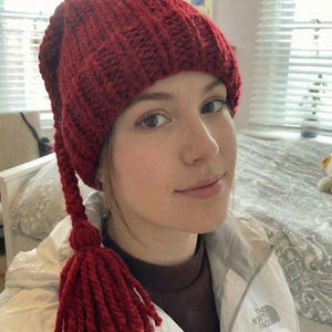 May include: A knitted red hat with a long tassel. The hat is made of a thick, textured yarn and has a ribbed design. The tassel is made of the same yarn and hangs down from the side of the hat. The hat is a winter accessory.