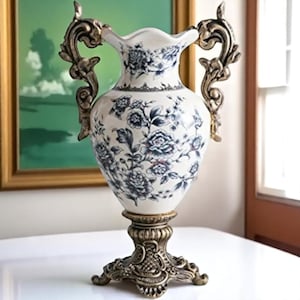 May include: A white ceramic vase with blue floral patterns and ornate bronze handles and base. The vase has a flared rim and is decorated with detailed blue flower and leaf designs, creating an elegant decorative piece.