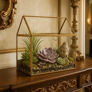 May include: A gold-framed terrarium with a house-like shape, showcasing a miniature garden. Inside, a variety of succulents, including a large lavender-colored succulent, are arranged with small pebbles and a Buddha statue.