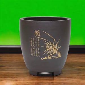 May include: A dark grey ceramic planter with a gold design of a plant and the Chinese character "蘭" (Lan). The planter has a wide base and a narrow opening.