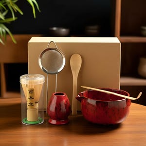 May include: A matcha tea set includes a red ceramic bowl, bamboo whisk, bamboo scoop, and a red ceramic whisk holder. A stainless steel tea strainer and a container with a bamboo whisk are also included. The set is displayed on a wooden surface.