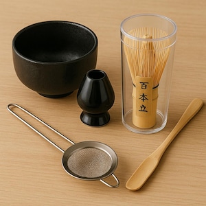 May include: A matcha tea set featuring a black ceramic bowl, a black ceramic whisk holder, a bamboo whisk in a clear case, a stainless steel sifter, and a wooden scoop. The items are arranged on a light wood surface.