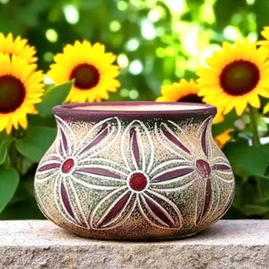 May include: A decorative ceramic flower pot with a beige and green background, featuring a floral design in burgundy and white. The pot is sitting on a stone surface, with sunflowers in the background.
