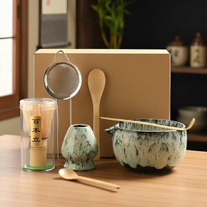 May include: A matcha tea set includes a ceramic bowl with a black and green glaze, a bamboo whisk, a bamboo scoop, a tea strainer, a whisk holder, and a wooden spoon. The set is presented on a wooden surface.