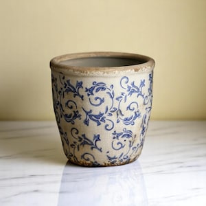 May include: A cylindrical ceramic plant pot with a distressed finish. The off-white pot has a blue floral pattern and a weathered rim. It sits on a white marble surface. The pot's design evokes a vintage aesthetic.