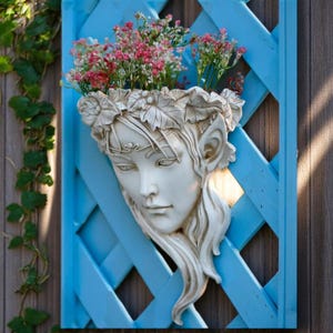 May include: A white face planter with a floral crown, mounted on a blue lattice. The planter holds pink and red flowers. The sculpture is a decorative garden feature, adding a touch of fantasy.