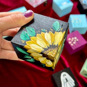 May include: A small, black wooden box with a hand-painted sunflower design in yellow and green. The box is held in a hand with purple nail polish. Other small, painted boxes in various colors are in the background.