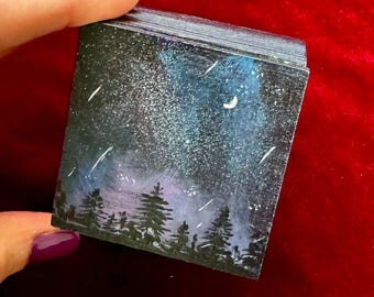 Può includere: Una piccola opera d'arte quadrata raffigurante una scena di cielo notturno con una silhouette di foresta. Il dipinto presenta uno sfondo blu scuro e nero con stelle bianche, una luna crescente e strisce. La foresta è resa in nero.