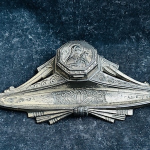 Peut inclure: Une pièce décorative antique en métal argenté avec un médaillon central octogonal représentant une figure religieuse. La pièce présente un design Art Déco avec des motifs floraux et géométriques stylisés.