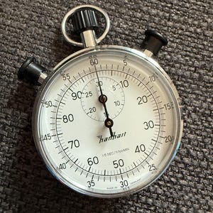 May include: A silver and black Hanhart stopwatch with a white face, featuring black numerals and markings. The stopwatch has a prominent crown and two push buttons. The face displays the text "Hanhart" and "Made in Germany".