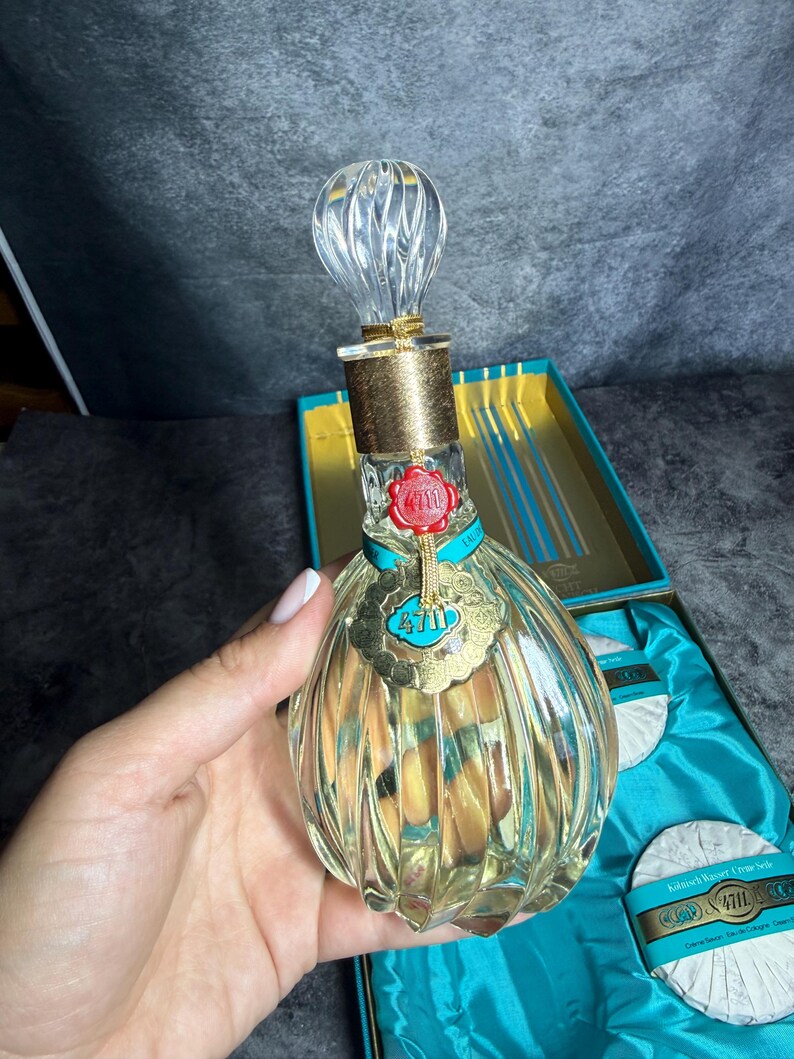 Puede incluir: Un frasco de perfume de vidrio transparente con dise&ntilde;o acanalado, cuello dorado y tap&oacute;n transparente retorcido. El frasco presenta un sello de cera roja y una etiqueta dorada con el texto 4711. Se presenta en una caja turquesa con dos jabones redondos.