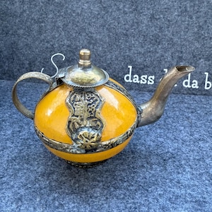 Könnte beinhalten: Eine Vintage-Teekanne mit einem gelben, rissigen Keramikkörper und verzierten Metallakzenten. Die Teekanne hat einen gebogenen Ausguss, einen Henkel und einen Deckel mit einem kleinen Knopf. Der Text "dass da b" ist auf der rechten Seite der Teekanne zu sehen.