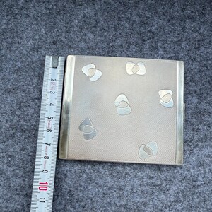 May include: A silver-toned cigarette case with a textured surface and decorative silver-colored shapes. The case is rectangular with a hinged opening and a metal border. A ruler is placed next to the case, showing measurements in centimeters.