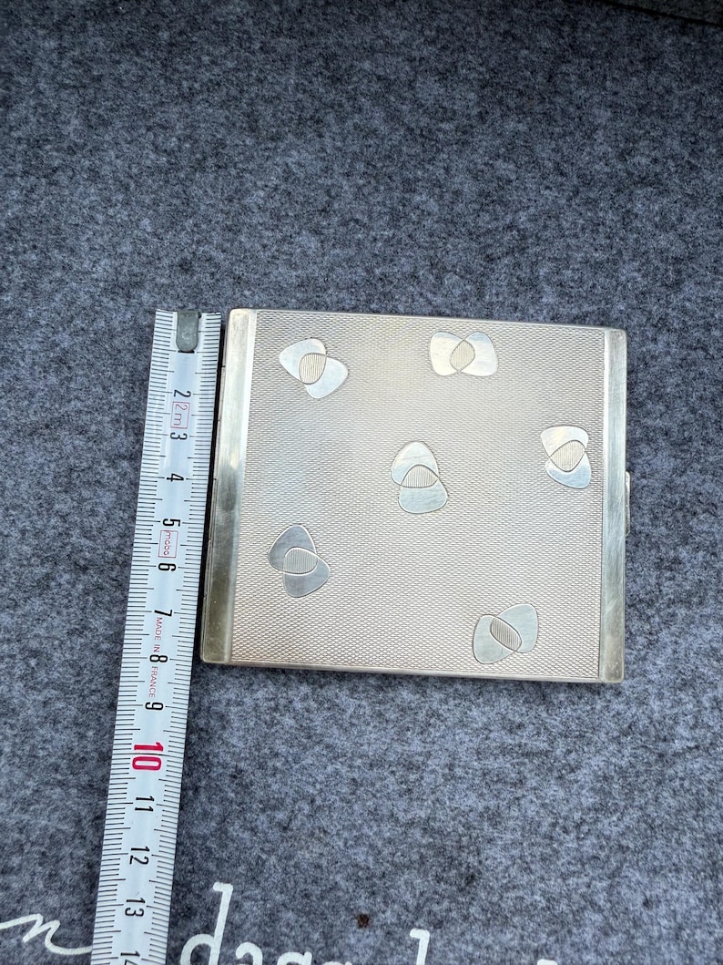 May include: A silver-toned cigarette case with a textured surface and decorative silver-colored shapes. The case is rectangular with a hinged opening and a metal border. A ruler is placed next to the case, showing measurements in centimeters.