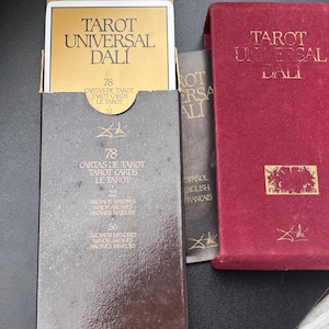 Pu&ograve; includere: Un set di carte Tarot Universal Dali in una scatola marrone con scritte dorate. La scatola &egrave; aperta, rivelando le carte e la parte superiore della scatola. &Egrave; visibile anche una scatola rossa con scritte dorate. Il testo sulla scatola recita "Tarot Universal Dali".