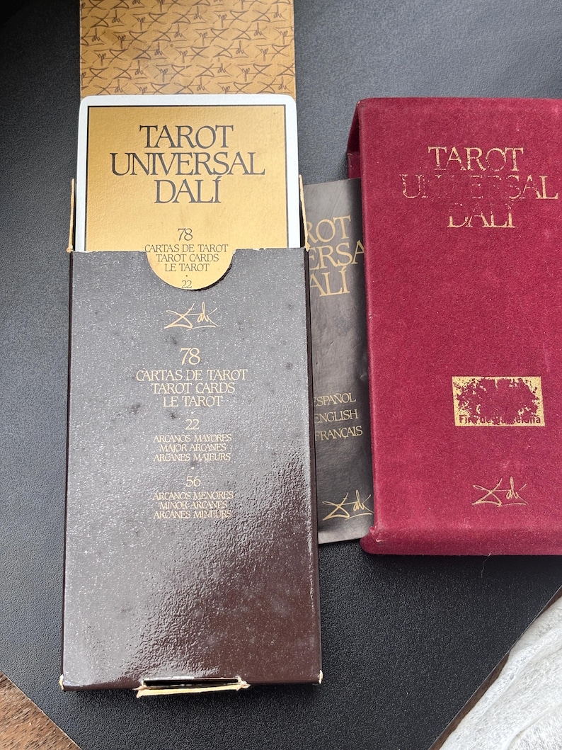 Pu&ograve; includere: Un set di carte Tarot Universal Dali in una scatola marrone con scritte dorate. La scatola &egrave; aperta, rivelando le carte e la parte superiore della scatola. &Egrave; visibile anche una scatola rossa con scritte dorate. Il testo sulla scatola recita "Tarot Universal Dali".