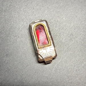 May include: A rectangular silver-toned metal reliquary featuring a central arched window framed by a textured gold-toned border. The interior displays a deep red fabric inlay. The lower portion of the metal casing is engraved with a sunburst pattern.