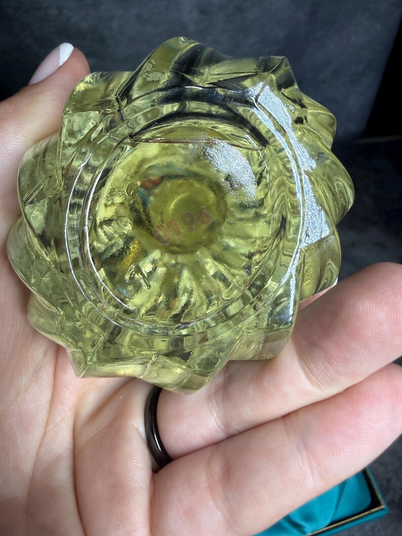 Puede incluir: Un objeto de vidrio transl&uacute;cido de color amarillo p&aacute;lido con una forma geom&eacute;trica facetada en forma de estrella. La base presenta una hendidura circular con el n&uacute;mero 0196 grabado en el centro. El vidrio tiene una superficie lisa y brillante.