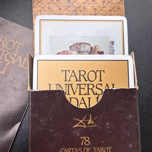 Pu&ograve; includere: Una scatola di carte dei tarocchi marrone con scritte dorate e le parole "TAROT UNIVERSAL DALI". La scatola contiene carte dei tarocchi con illustrazioni. La scatola ha anche il numero "78" e la firma dell'artista.