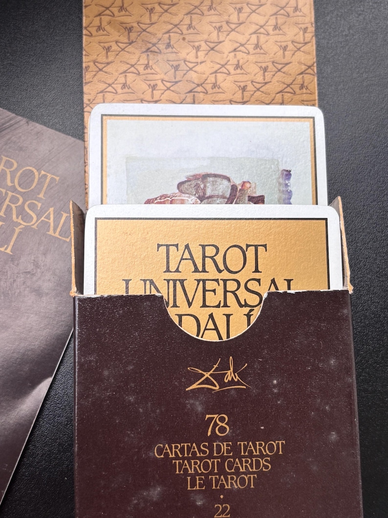 Pu&ograve; includere: Una scatola di carte dei tarocchi marrone con scritte dorate e le parole "TAROT UNIVERSAL DALI". La scatola contiene carte dei tarocchi con illustrazioni. La scatola ha anche il numero "78" e la firma dell'artista.