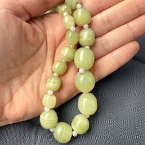 Könnte beinhalten: Eine Halskette mit hellgrünen, oval geformten Perlen mit feinen Streifen, die durch kleine weiße Perlen getrennt sind. Die Kette wird vor einem grauen Hintergrund präsentiert.