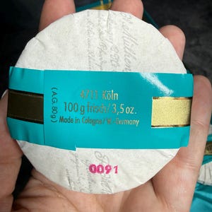 Puede incluir: Una pastilla de jab&oacute;n redonda 4711 K&ouml;ln envuelta en papel blanco con una banda turquesa. La banda presenta texto dorado que dice 4711 K&ouml;ln, 100 g frisch/3,5 oz. y Made in Cologne/W.-Germany. El n&uacute;mero 0091 est&aacute; impreso en rosa sobre el papel.