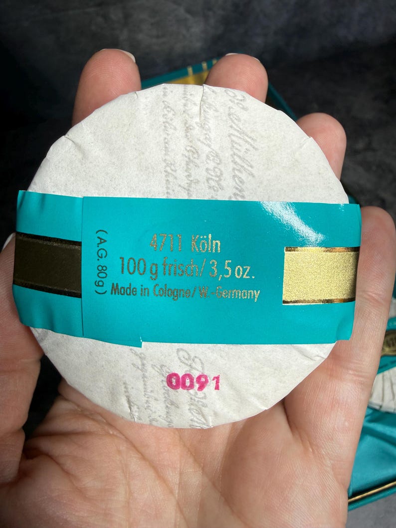 Puede incluir: Una pastilla de jab&oacute;n redonda 4711 K&ouml;ln envuelta en papel blanco con una banda turquesa. La banda presenta texto dorado que dice 4711 K&ouml;ln, 100 g frisch/3,5 oz. y Made in Cologne/W.-Germany. El n&uacute;mero 0091 est&aacute; impreso en rosa sobre el papel.