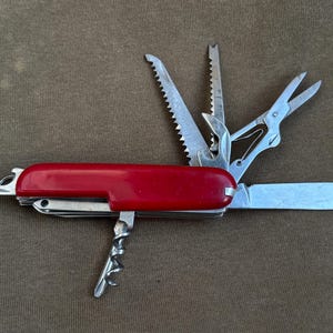 Könnte beinhalten: Ein rotes Multitool mit verschiedenen Werkzeugen, darunter ein Messer, eine Schere und Sägen. Das Werkzeug liegt auf einer braunen Oberfläche. Die Klingenlänge des Messers beträgt ca. 7,6 cm.