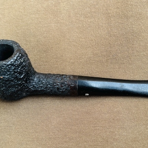 May include: A black tobacco pipe with a textured bowl and a smooth, dark stem. The pipe is made of a dark material and has a classic shape. The pipe is a smoking accessory.
