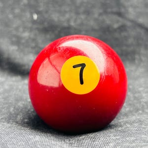 Puede incluir: Una bola de billar roja brillante con un círculo amarillo que muestra el número 7 en negro. La bola es esférica y el fondo es una tela gris texturizada.