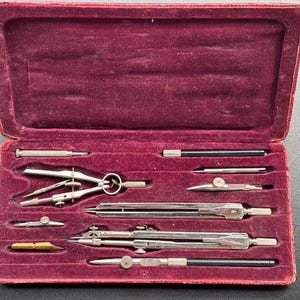 Könnte beinhalten: Ein offener, bordeauxroter, samtgefütterter Koffer mit einem Satz Vintage-Zeichenwerkzeuge. Das Set enthält mehrere silberfarbene Zirkel, Teiler und andere Präzisionsinstrumente sowie einen schwarzen Stift und einen kleinen Schraubendreher.