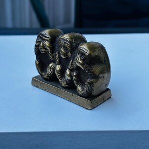 May include: Three small, gold-colored monkey figurines, each covering a different part of their face, are arranged in a row on a small, rectangular base. The monkeys are sitting with their legs crossed and their hands covering their eyes, ears, and mouth.