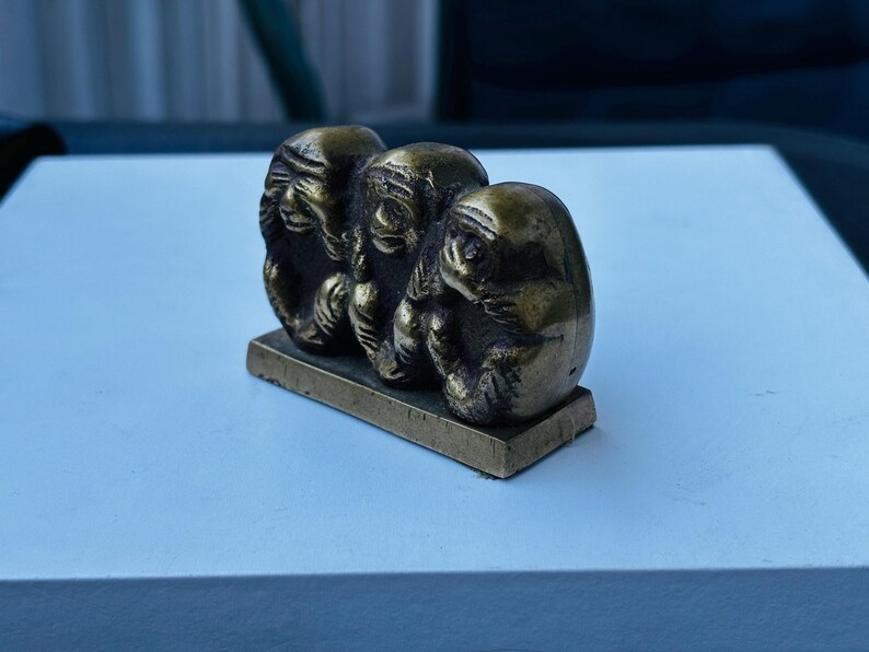May include: Three small, gold-colored monkey figurines, each covering a different part of their face, are arranged in a row on a small, rectangular base. The monkeys are sitting with their legs crossed and their hands covering their eyes, ears, and mouth.