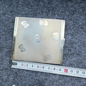 May include: A silver metal cigarette case with a textured surface and abstract designs. The case is rectangular and measures approximately 3.5 inches in length. A ruler is placed next to the case.