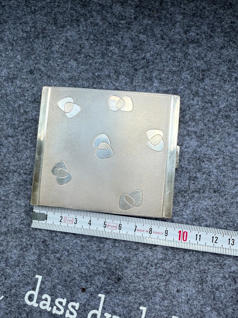May include: A silver metal cigarette case with a textured surface and abstract designs. The case is rectangular and measures approximately 3.5 inches in length. A ruler is placed next to the case.