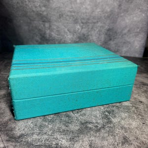 Puede incluir: Una caja rectangular con un exterior de tela texturizada en color verde azulado. La tapa presenta tres finas rayas horizontales de tono dorado que atraviesan la superficie superior. La caja tiene un dise&ntilde;o estructurado con bordes definidos.
