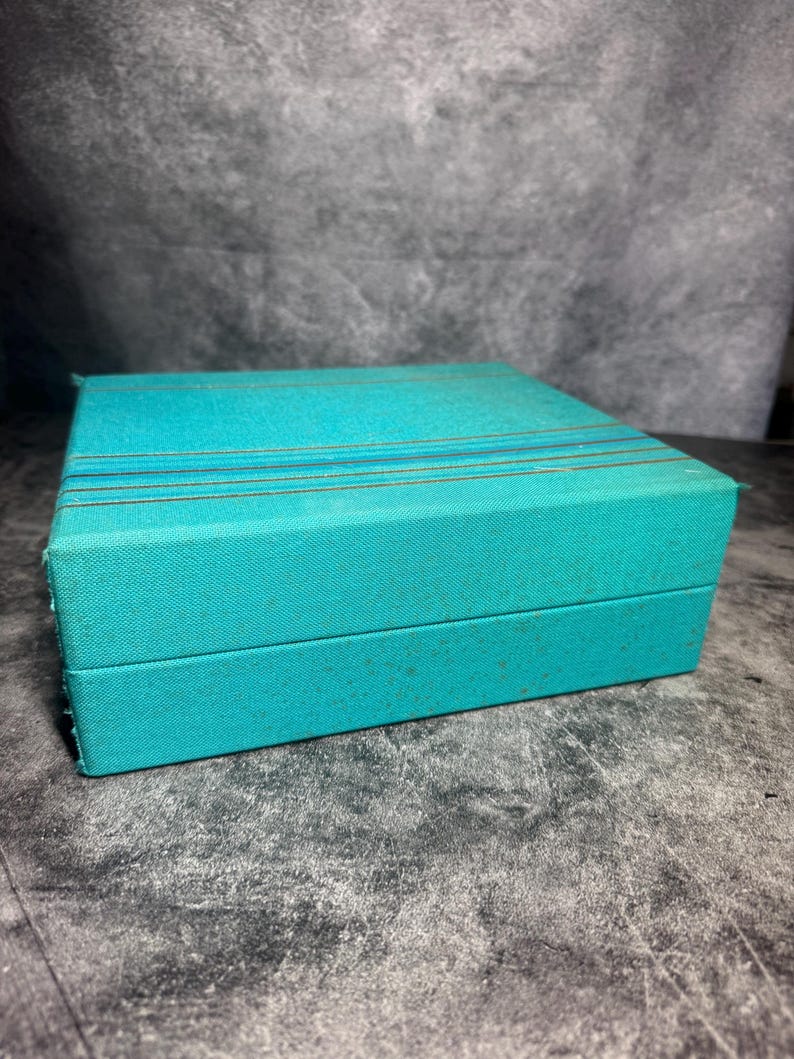 Puede incluir: Una caja rectangular con un exterior de tela texturizada en color verde azulado. La tapa presenta tres finas rayas horizontales de tono dorado que atraviesan la superficie superior. La caja tiene un dise&ntilde;o estructurado con bordes definidos.