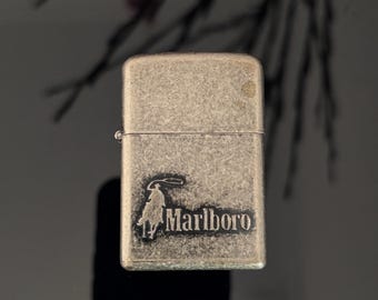 Puede incluir: Un encendedor plateado vintage con el logotipo de Marlboro, que presenta una silueta de vaquero y el nombre de la marca. El encendedor tiene forma rectangular y una tapa con bisagras. La superficie muestra signos de desgaste.