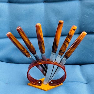 Può includere: Un set di sei coltelli da bistecca con lame in acciaio inossidabile e manici color ambra. I coltelli sono esposti in un supporto abbinato con una base arancione e un design curvo ad anello. I manici hanno un aspetto marmorizzato.