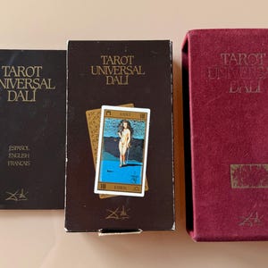 Pu&ograve; includere: Un set di Tarot Universal Dali. Include un libro nero con testi in spagnolo, inglese e francese, una scatola con una carta e una scatola bordeaux. Tutti gli articoli riportano il nome "Tarot Universal Dali".