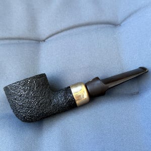 May include: A black tobacco pipe with a textured bowl and a dark brown stem. The pipe has a gold-coloured band near the stem. The pipe is designed for smoking tobacco.