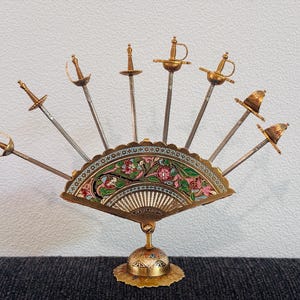 Może przedstawiać: Ozdobny przedmiot w kształcie wachlarza z miniaturowymi mieczami. Wachlarz ma złotą ramę z emaliowanym kwiatowym wzorem w kolorze różowym, zielonym i białym. Miecze są srebrne ze złotymi rękojeściami i są ułożone tak, aby przypominały kształt wachlarza.