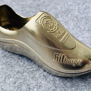 Op de afbeelding: Een goudkleurige flesopener in de vorm van een schoen met het woord "Bitburger" in reliëf aan de zijkant. De opener heeft een logo aan de bovenkant en is gemaakt van glanzend metaal.