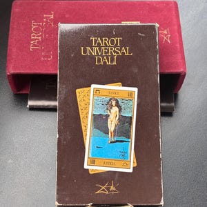Pu&ograve; includere: Una scatola di carte Tarot Universal Dali marrone con la scritta "Tarot Universal Dali" in lettere dorate. La scatola presenta una carta con l'illustrazione di una figura nuda che tiene una spada. Dietro c'&egrave; una scatola di velluto rosso.