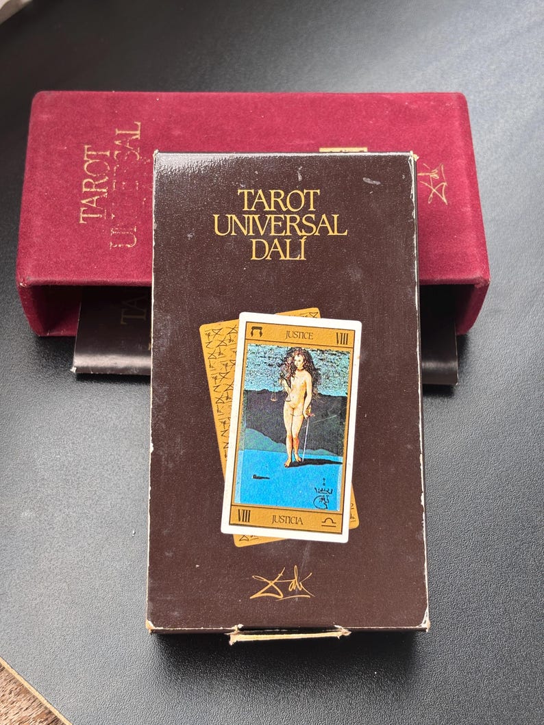 Pu&ograve; includere: Una scatola di carte Tarot Universal Dali marrone con la scritta "Tarot Universal Dali" in lettere dorate. La scatola presenta una carta con l'illustrazione di una figura nuda che tiene una spada. Dietro c'&egrave; una scatola di velluto rosso.