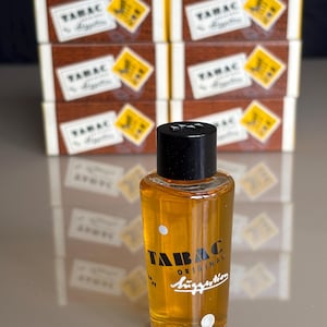 Può includere: Flacone in vetro trasparente di dopobarba Tabac Original con tappo nero. La bottiglia contiene un liquido ambrato e scritte bianche. Dietro la bottiglia ci sono diverse scatole con la scritta "TAMAC" e un disegno giallo e bianco.