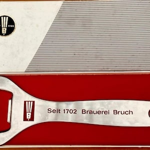 May include: A silver bottle opener with a red and white logo that says "Seit 1702 Brauerei Bruch". The bottle opener is in a red velvet box.