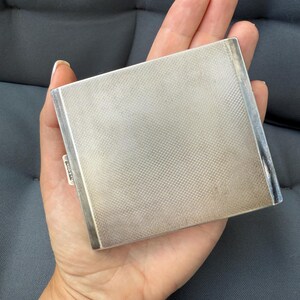 May include: A silver-colored, rectangular compact with a textured, cross-hatched surface. The compact has a simple, elegant design and a small, rectangular clasp. The item is held in a hand.