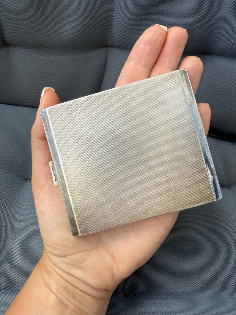 May include: A silver-colored, rectangular compact with a textured, cross-hatched surface. The compact has a simple, elegant design and a small, rectangular clasp. The item is held in a hand.