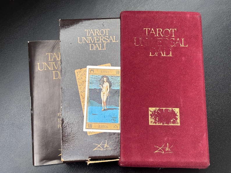 Pu&ograve; includere: Una collezione di mazzi di Tarocchi Universal Dali. I mazzi sono in una variet&agrave; di colori, tra cui marrone scuro, grigio scuro e rosso bordeaux. I mazzi hanno scritte dorate e presentano una carta con un'illustrazione di una donna nuda.
